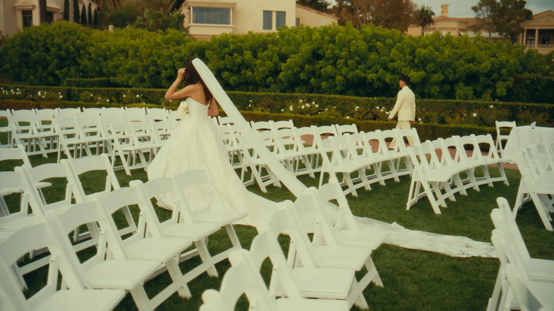 A bride in a long veil stands among rows of white ceremony chairs as the groom walks toward her