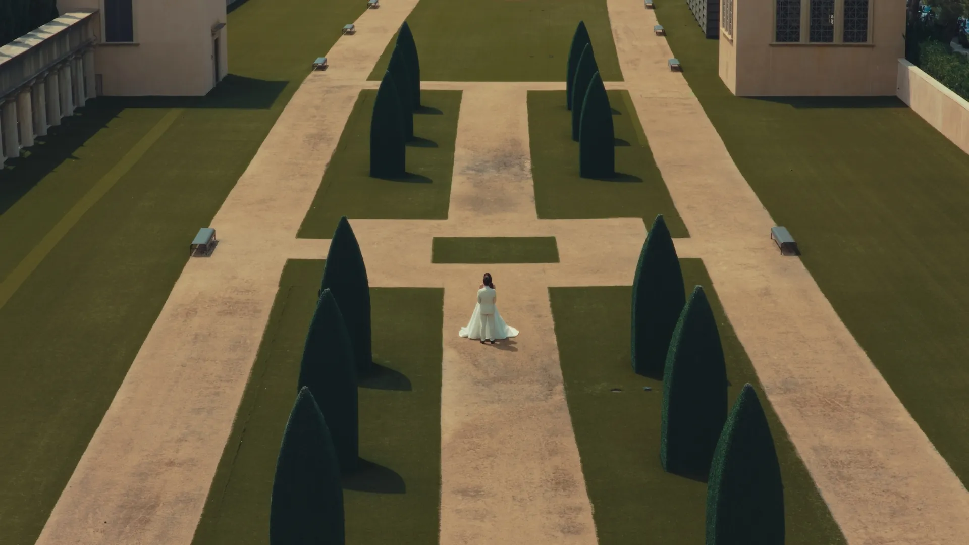 Aerial view of a bride and groom standing on a gravel path between rows of topiary